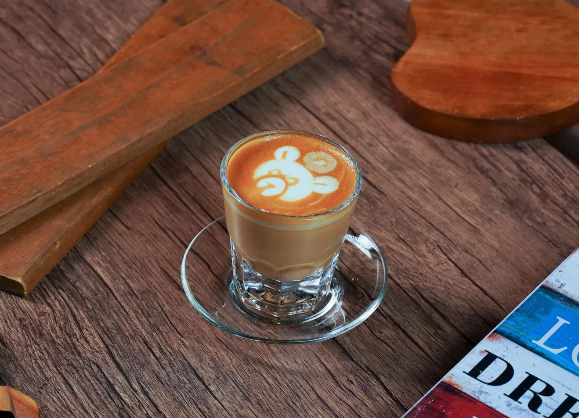 Glass cup of Turkish coffee with bear latte art on rustic wooden table beside wooden coasters and colorful book