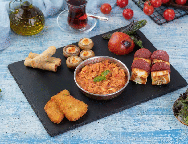 A black serving board displaying a variety of Turkish appetizers, including a bowl of scrambled-style tomato and egg mixture, fried rolls, stuffed mushrooms, fried cheese pieces topped with sausage slices, and crispy breaded bites. Fresh vegetables, olive oil, and a glass of tea are arranged around the board on a blue wooden surface.