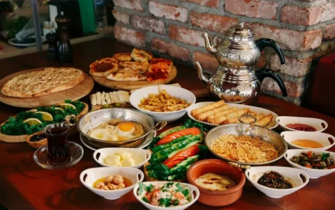 A large traditional Turkish breakfast spread featuring fried eggs, borek rolls, fries, fresh vegetables, cheeses, various dips and sauces, pastries, flatbreads, and Turkish tea, arranged on a wooden table with a decorative double teapot in the background.