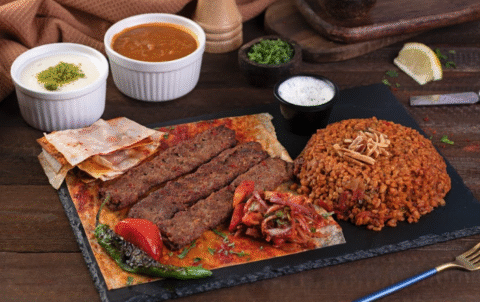 A Turkish meal served on a black slate plate, featuring grilled minced meat kebabs on flatbread, a mound of bulgur pilaf topped with slivered nuts, roasted vegetables, and a side of tomato-onion salad. Small bowls of yogurt, soup, and sauce are placed in the background on a wooden table.