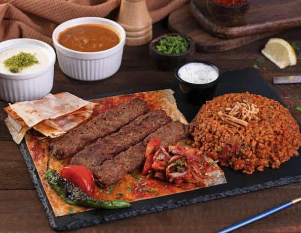 A Turkish meal served on a black slate plate, featuring grilled minced meat kebabs on flatbread, a mound of bulgur pilaf topped with slivered nuts, roasted vegetables, and a side of tomato-onion salad. Small bowls of yogurt, soup, and sauce are placed in the background on a wooden table.