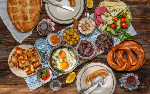 A beautifully arranged Turkish breakfast spread on a wooden table featuring fried eggs with herbs, assorted cheeses, cold cuts, olives, fresh vegetables, jam, honey, dates, simit bread rings, pide bread, börek pastries, and glasses of Turkish tea.