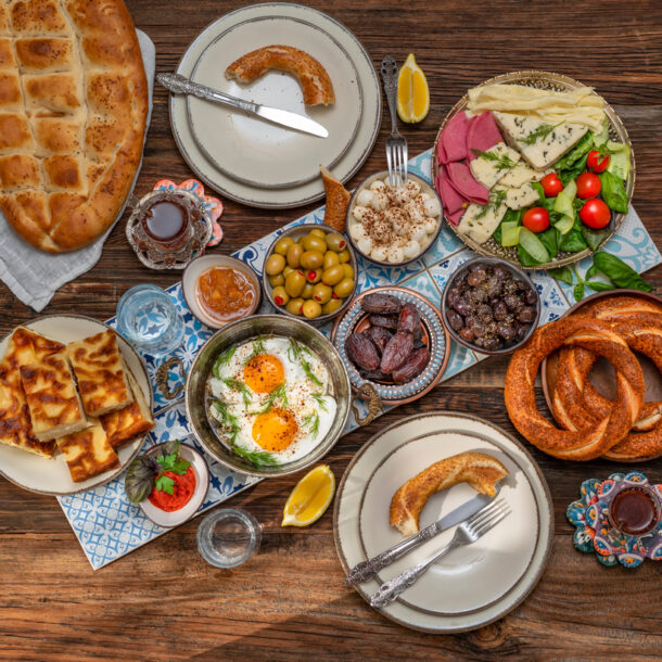 A beautifully arranged Turkish breakfast spread on a wooden table featuring fried eggs with herbs, assorted cheeses, cold cuts, olives, fresh vegetables, jam, honey, dates, simit bread rings, pide bread, börek pastries, and glasses of Turkish tea.