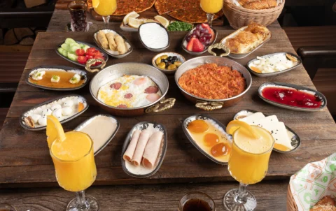 Family-style Turkish breakfast spread at a Turkish restaurant in Abu Dhabi, featuring freshly baked pide, eggs, cheeses, olives, pastries, grilled dishes, tea, and fresh juice served on a wooden table in a warm dining setting.