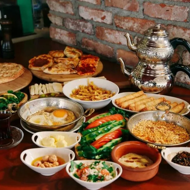 A large traditional Turkish breakfast spread featuring fried eggs, borek rolls, fries, fresh vegetables, cheeses, various dips and sauces, pastries, flatbreads, and Turkish tea, arranged on a wooden table with a decorative double teapot in the background.
